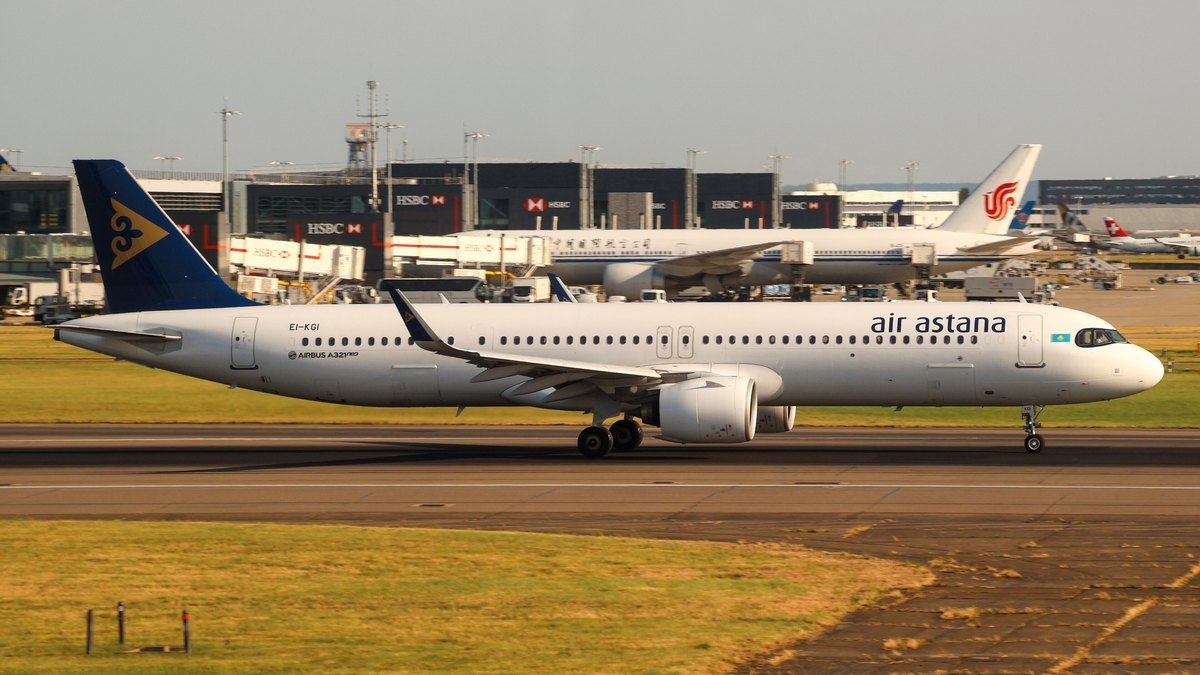 Air Astana Airbus A321neo EI-KGI on the runway at London Heathrow in June 2025, white with a dark blue tail carrying a gold emblem, with terminal buildings and an Air China jet behind.