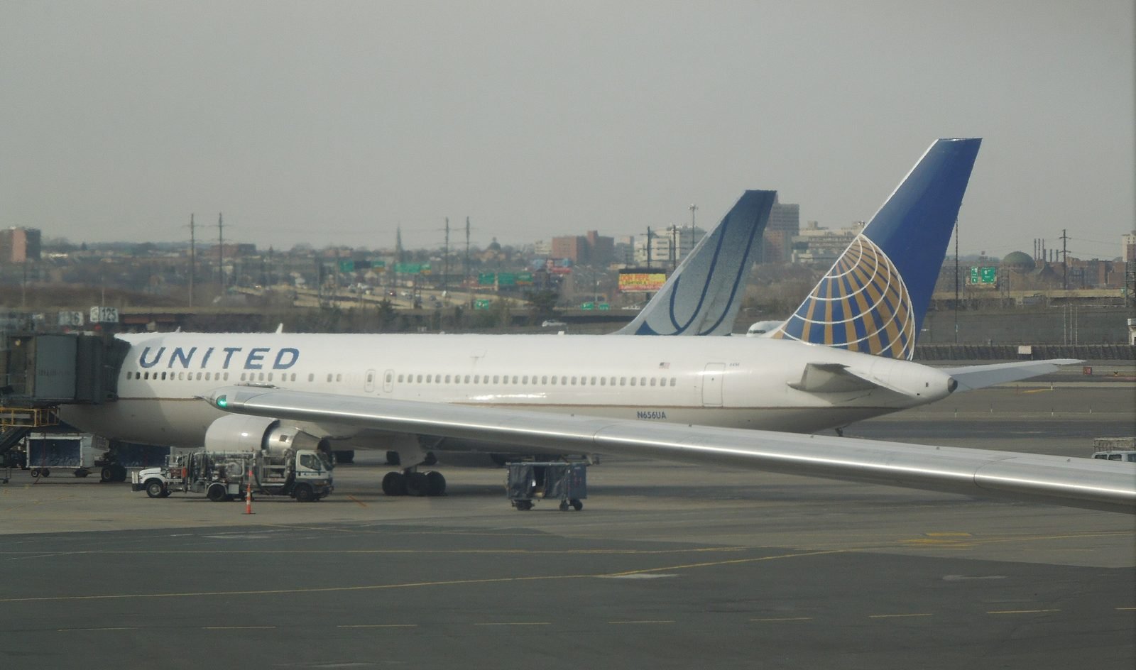 United Boeing 767-300ER N656UA parked at a gate at Newark in the globe livery, seen through a terminal window with another jet's wing in the foreground.