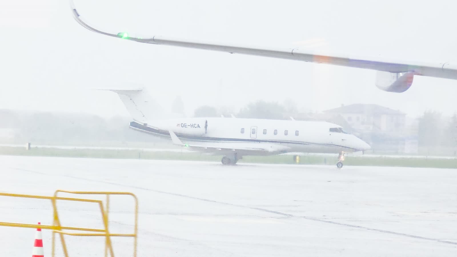 A white Bombardier Challenger 300 business jet, registration OE-HCA, parked on a rain-soaked apron at Verona airport in grey, misty weather.