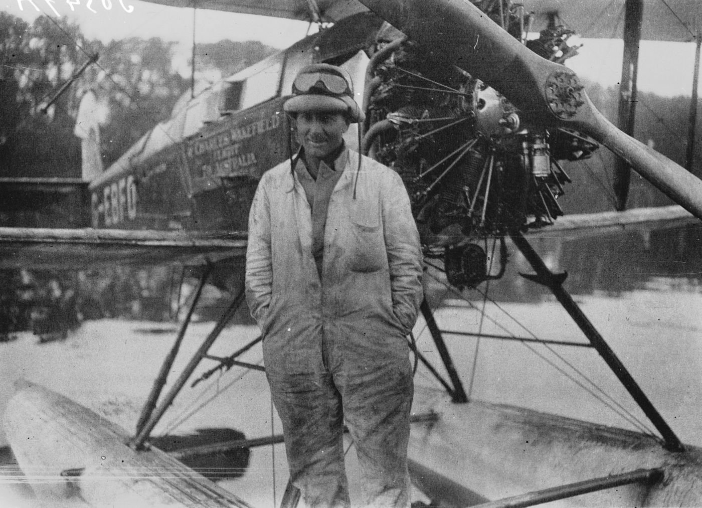 Black and white photograph of Alan Cobham in flying overalls and cap standing on the float of his DH.50 floatplane, its engine and propeller behind him.
