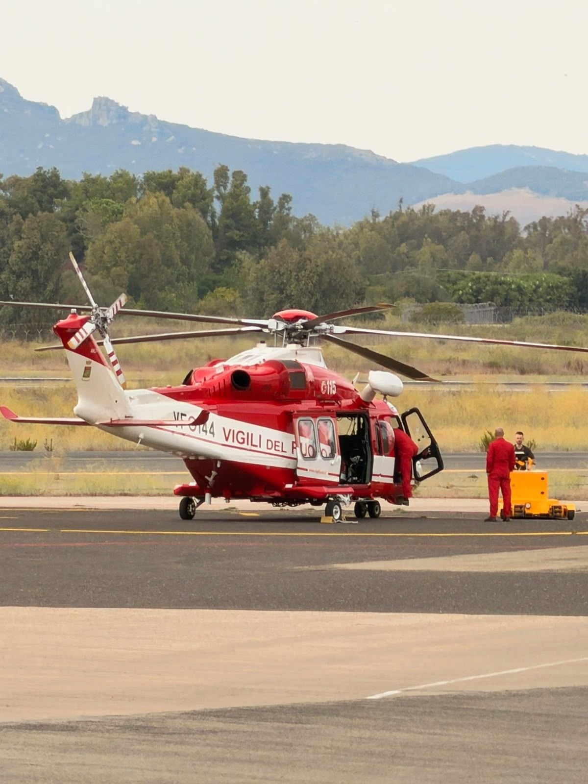 A red and white Leonardo AW139 helicopter of Italy's Vigili del Fuoco on the ground in Sardinia, with 115 on its nose, VF-144 on the tail boom and two crew in red suits at the open door. Photo by a friend of the author.