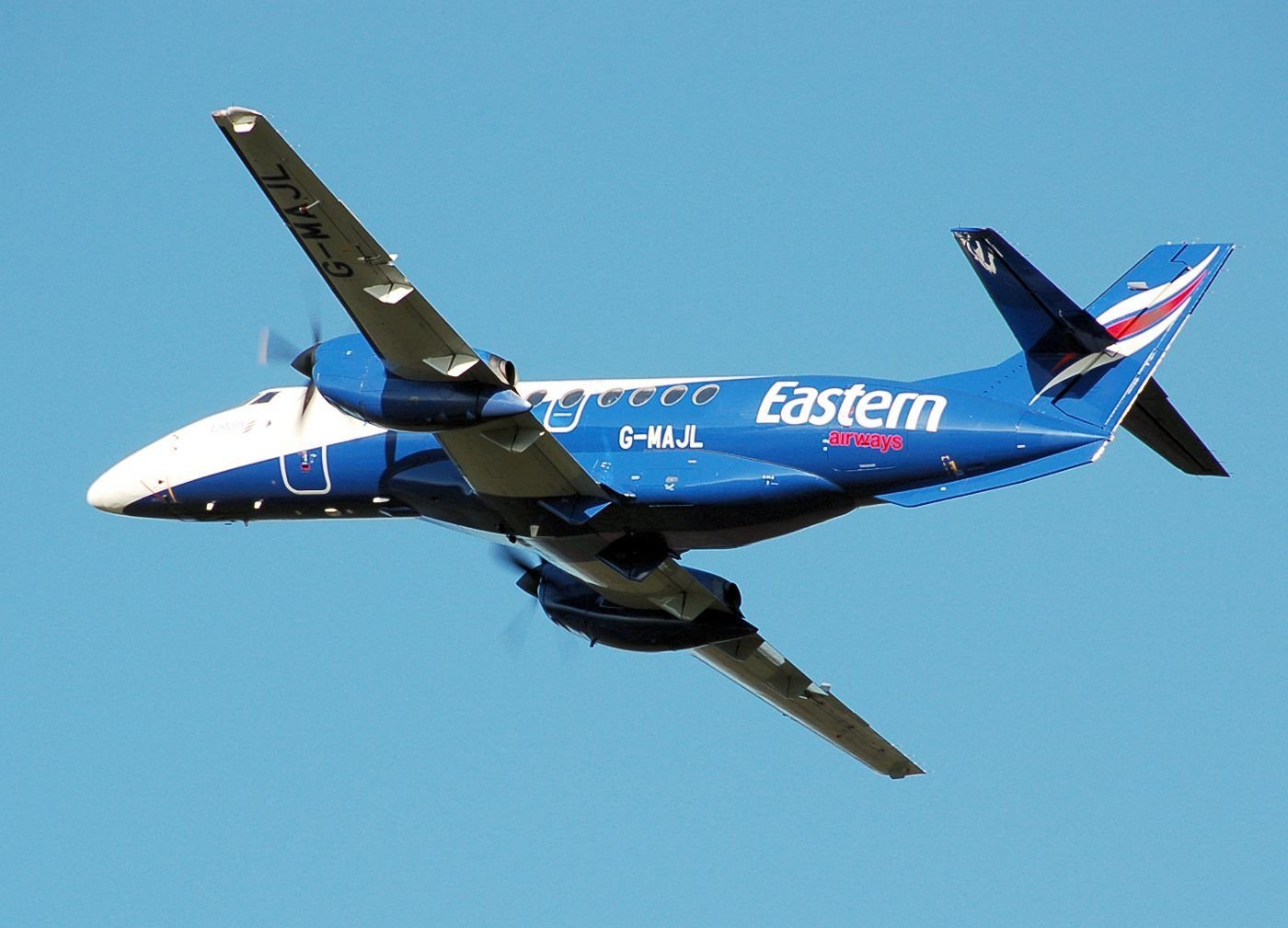 A blue and white Eastern Airways Jetstream 41 climbing away overhead, seen from below against a clear blue sky.