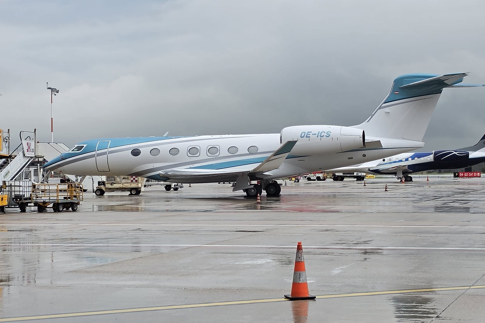 Gulfstream G600 registered OE-ICS parked side-on on a wet apron at Verona under low grey cloud, with its row of seven oval cabin windows clearly visible.