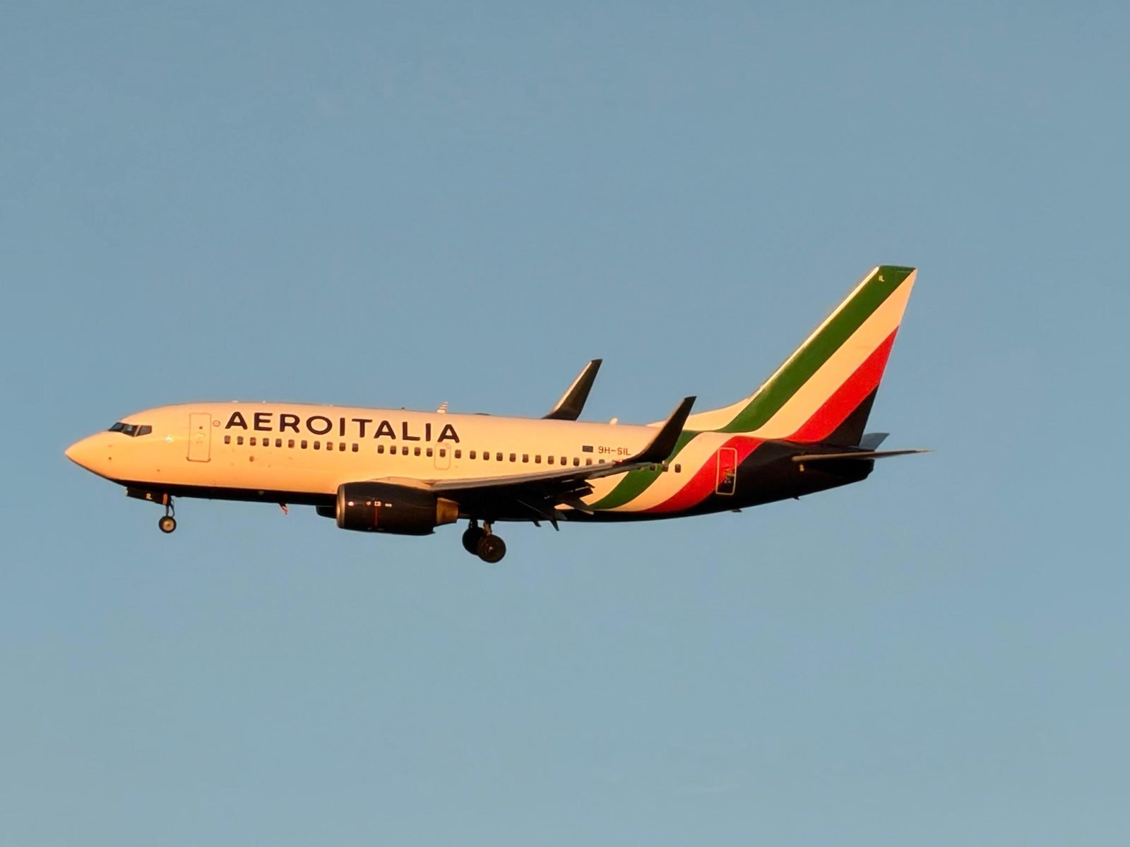 An Aeroitalia Boeing 737-700, registration 9H-SIL, on approach with its gear down, lit gold by the evening sun.